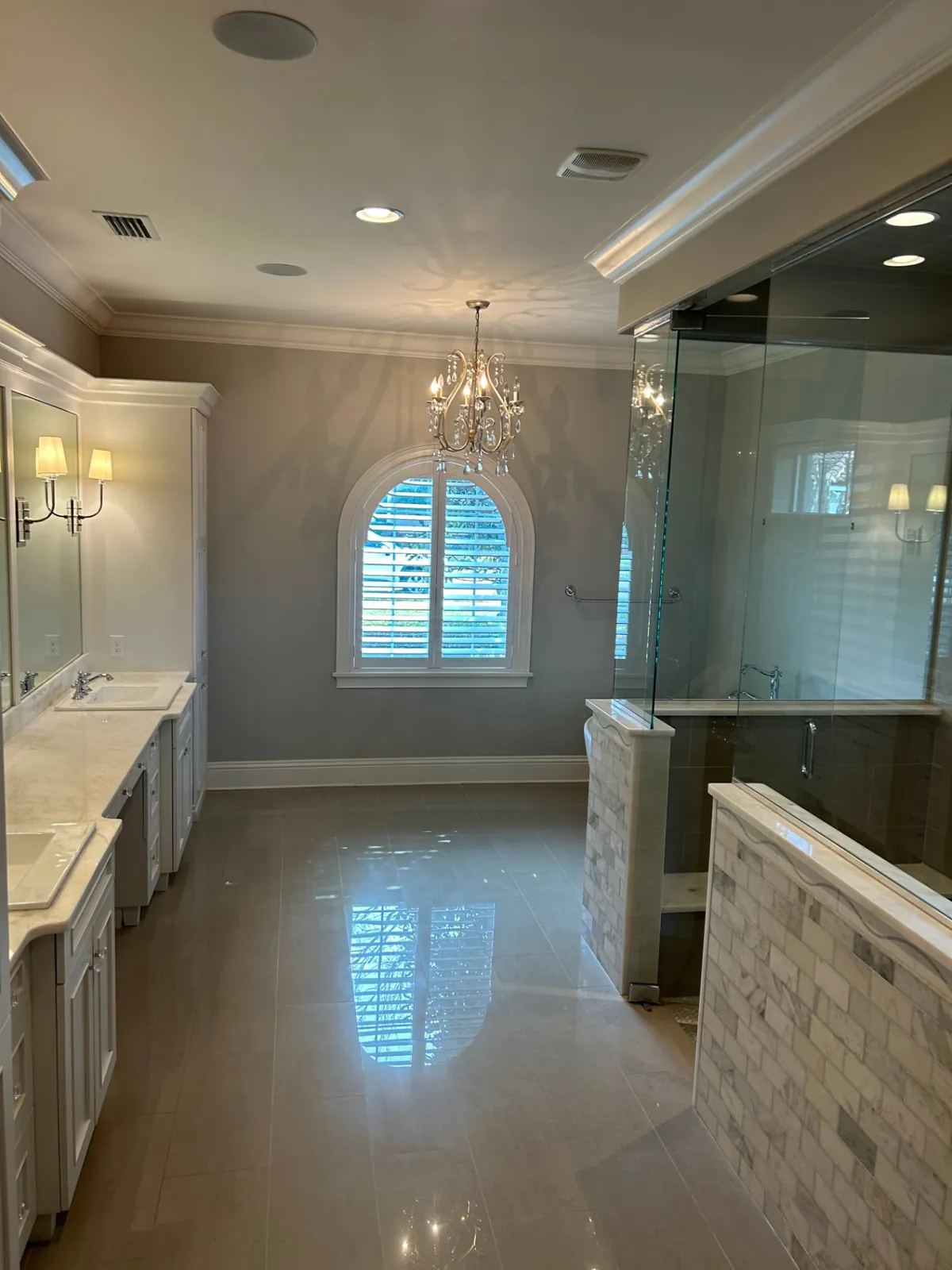 Finished: Primary bathroom with white vanities, crystal chandelier, arched window and a frameless glass shower